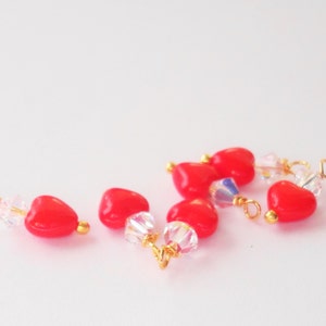 May include: A group of red heart-shaped beads with gold accents and clear crystal beads. The beads are scattered on a white surface.