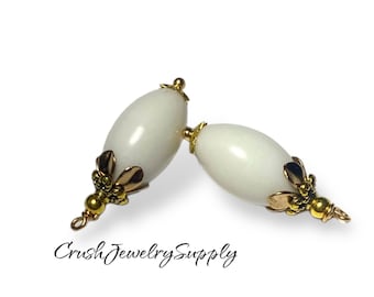 May include: Two white oval-shaped beads with gold accents. The beads have a loop at the bottom for attaching to jewelry.
