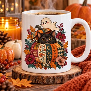 Op de afbeelding: Een witte keramische mok met een kleurrijke illustratie van een zwarte kat die uit een pompoen met patronen kijkt, bekroond met een klein spookje, omringd door herfstbladeren, bessen en bloemen, met stoom die uit de koffie opstijgt.