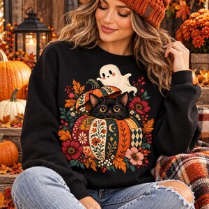 Op de afbeelding: Een zwart sweatshirt met lange mouwen en een grafische print van een zwarte kat die uit een pompoen met patronen kijkt, omringd door herfstbladeren, bloemen en een klein wit spookje dat boven de pompoen zweeft, tegen een donkere achtergrond.