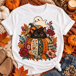 Peut inclure: Un t-shirt blanc avec une impression graphique représentant un chat noir sortant d'une citrouille à motifs, surmonté d'un petit fantôme. Le motif est entouré de feuilles d'automne colorées, de baies rouges et de fleurs aux tons rouges et roses.