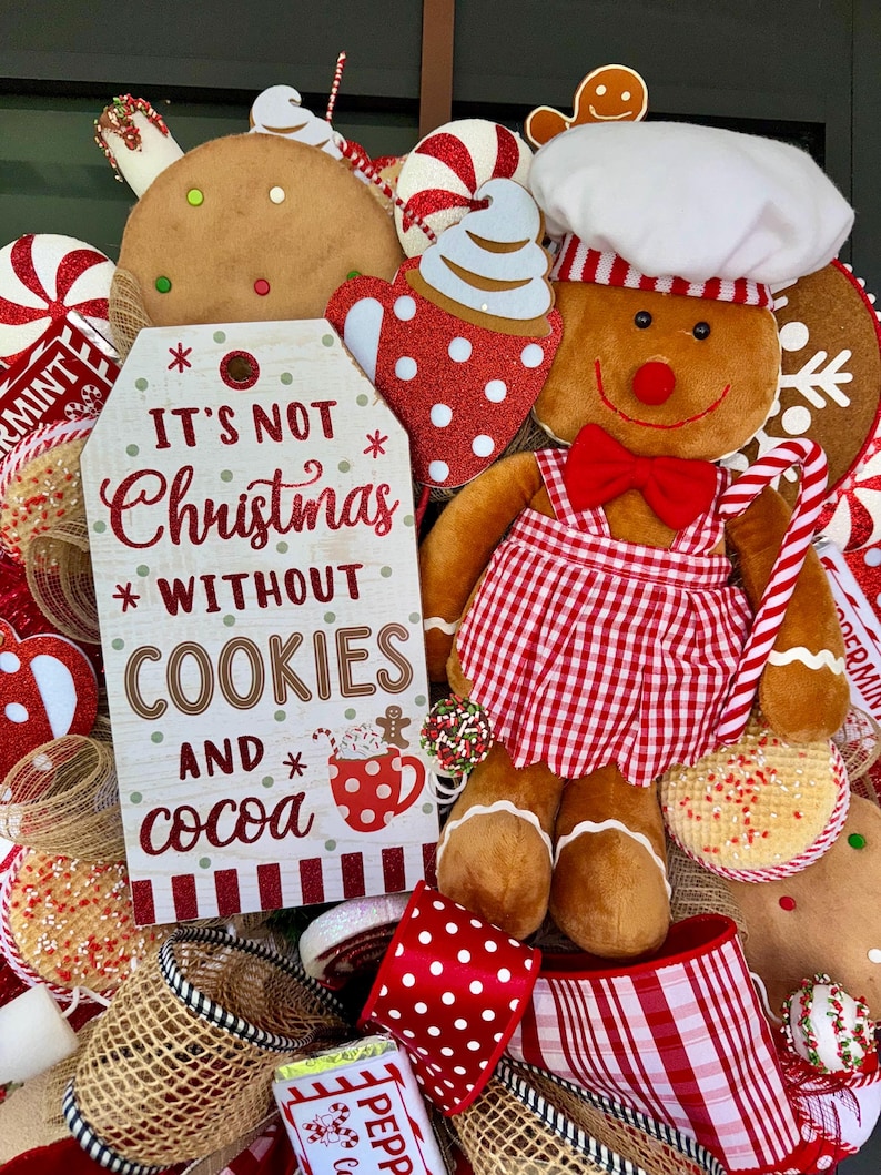 Puede incluir: Una corona navide&ntilde;a festiva con un hombre de jengibre con gorro y delantal de chef, sosteniendo un bast&oacute;n de caramelo. La corona incluye galletas de jengibre, mentas y un letrero que dice "It's not Christmas without cookies and cocoa."