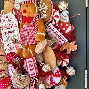 Puede incluir: Una corona navide&ntilde;a festiva con un hombre de jengibre chef, galletas y caramelos de menta. La corona incluye un letrero que dice "It's not Christmas without cookies and cocoa". Los colores dominantes son el rojo, el blanco y el marr&oacute;n.