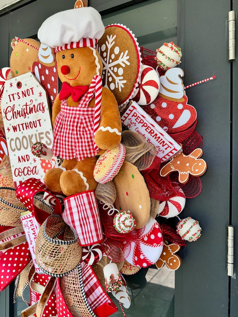 Puede incluir: Una corona navide&ntilde;a festiva con un hombre de jengibre chef, galletas y caramelos de menta. La corona incluye un letrero que dice "It's not Christmas without cookies and cocoa". Los colores dominantes son el rojo, el blanco y el marr&oacute;n.