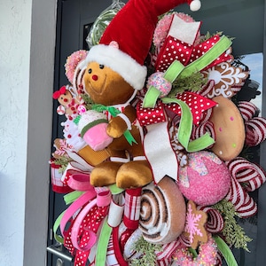 Puede incluir: Una corona navide&ntilde;a festiva con un hombre de jengibre con un gorro de Pap&aacute; Noel. La corona est&aacute; adornada con cintas coloridas, productos horneados falsos y elementos decorativos. La paleta de colores incluye rojo, verde, rosa y blanco.