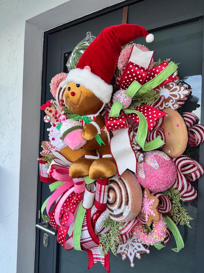Puede incluir: Una corona navide&ntilde;a festiva con un hombre de jengibre con un gorro de Pap&aacute; Noel. La corona est&aacute; adornada con cintas coloridas, productos horneados falsos y elementos decorativos. La paleta de colores incluye rojo, verde, rosa y blanco.