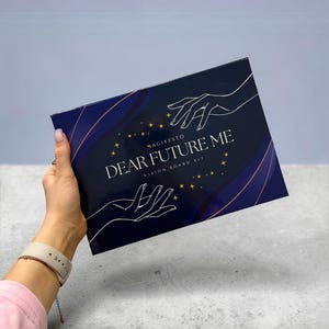 Könnte beinhalten: Ein dunkelblaues Vision Board Kit mit dem Titel "DEAR FUTURE ME" mit weißem Text und Sternakzenten. Die Box zeigt ein Design von sich ausstreckenden Händen und das Wort "MAGIFESTO". Das Kit wird von einer Person gehalten.