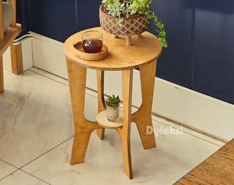 Könnte beinhalten: Ein hellbrauner Beistelltisch aus Holz mit runder Tischplatte und drei geschwungenen Beinen. Der Tisch verfügt über eine kleine untere Ablage, einen runden Holzuntersetzer mit einem Glas dunkler Flüssigkeit und einen gemusterten Topf mit Sukkulenten.