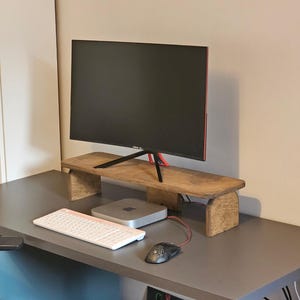 Puede incluir: Un soporte de monitor de madera con un monitor encima. Un teclado, un ratón y un ordenador están en el escritorio. El monitor tiene una pantalla negra y un borde rojo. El soporte es de madera marrón claro.