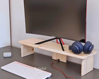Könnte beinhalten: Ein Monitorständer aus Naturholz mit rechteckiger Oberseite und zwei Stützbeinen. Auf dem Ständer steht ein schwarzer Computermonitor mit rotem Akzent. Eine weiße Tastatur, Maus und Kopfhörer sind ebenfalls sichtbar.