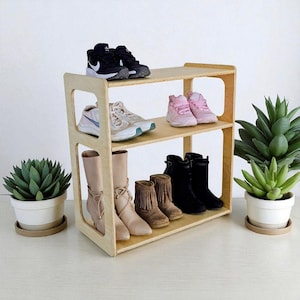 May include: A three-tiered, light-colored wooden shoe rack displaying various footwear. The top shelf holds black sneakers, the middle shelf has sneakers and pink shoes, and the bottom shelf features boots in beige, brown, and black. Potted succulents flank the rack.