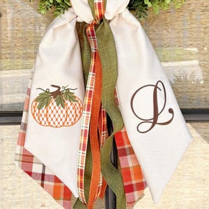 May include: A seasonal door decoration with a green wreath and cream fabric panels. The panels are adorned with an embroidered pumpkin and a letter 'D'. Ribbons in orange, green, and red complete the design.
