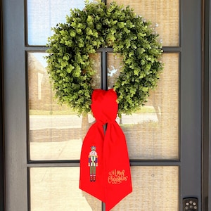 Puede incluir: Una corona verde con una cinta roja con una ilustración de cascanueces y texto dorado "Merry Christmas". La corona está hecha de pequeñas hojas verdes redondas y cuelga de una puerta de vidrio.