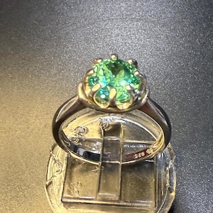 May include: A silver ring with a large, round, green gemstone. The gemstone is set in a decorative, gold-colored setting. The ring band is thin and silver. The ring is displayed on a small stand.