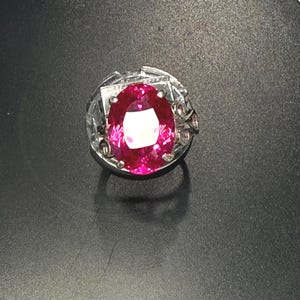 May include: A silver ring featuring a large, oval-cut, vibrant pink gemstone. The gemstone is the focal point, set in a detailed, ornate silver band with additional smaller pink stones. The ring is displayed on a dark surface.
