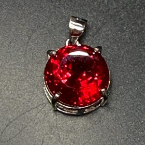 May include: A round, vibrant red gemstone pendant with a silver-colored setting. The gemstone is faceted, catching the light and creating a sparkling effect. The pendant has a simple, elegant design, suitable for everyday wear or special occasions.