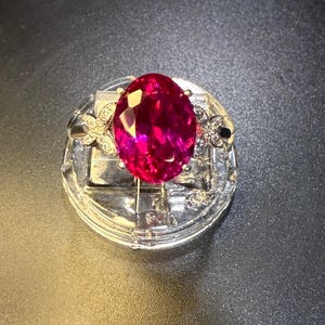 May include: A silver ring featuring a large, oval-cut, vibrant pink gemstone. The ring is accented with small, clear gemstones and floral designs on either side of the main stone. The ring is displayed on a clear, round stand.