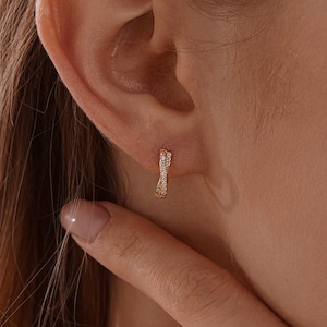 以下が含まれることがあります： 小さなクリアな宝石がセットされた、リボンのデザインのゴールドカラーのイヤリングのクローズアップ。イヤリングは人の耳に装着されています。背景は柔らかくニュートラルな色調です。