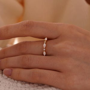 May include: A delicate gold ring featuring three oval-shaped gemstones and small accent stones. The ring is set on a hand, showcasing its elegant design and craftsmanship. The ring's design is simple yet sophisticated, perfect for everyday wear.