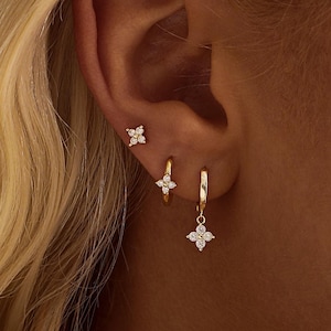 Könnte beinhalten: Goldenes Ohrring-Set mit einem kleinen Ohrstecker, einem Creolen-Ohrring mit Blumenanhänger und einem hängenden Blumenohrring. Die Ohrringe sind mit kleinen, funkelnden Steinen besetzt.