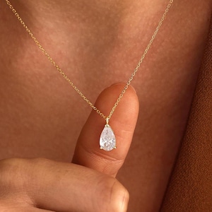 Puede incluir: Un delicado collar de cadena dorada con un colgante de piedra preciosa transparente en forma de pera. El colgante cuelga de una fina cadena de eslabones dorados pulidos que descansa sobre la piel, mostrando el corte facetado de la piedra.
