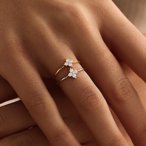 May include: Two rings, one gold and one silver, each featuring a cluster of small, sparkling gemstones in a floral design. The rings are displayed on a hand, highlighting the jewellery's intricate design and elegance. The gemstones are likely diamonds.