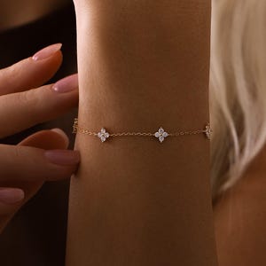 Könnte beinhalten: Ein zartes goldenes Armband mit vierblättrigen Kleeblatt-Designs mit kleinen, funkelnden Edelsteinen. Das Armband wird am Handgelenk getragen, eine Hand berührt es sanft. Das Armband ist ein feines Schmuckstück.