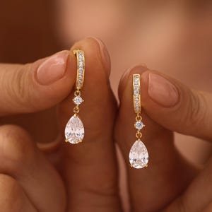 May include: A pair of gold hoop earrings featuring a small round clear stone and a dangling pear-shaped clear stone. The hoop is embellished with a row of tiny clear stones. The text LustreBloomDesign is visible in the bottom right corner.
