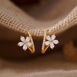 May include: A pair of gold hoop earrings featuring a flower-shaped charm adorned with clear, marquise-cut stones. Each hoop is accented with a row of small, round, clear stones along the outer edge. The text LustreBloomDesign is visible.
