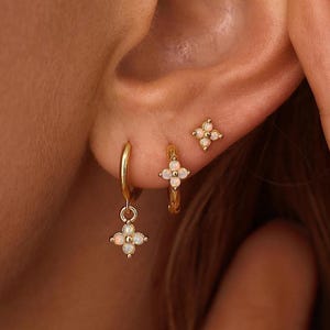 May include: Three gold earrings featuring a four-petaled flower design with small white opalescent stones. The set includes one small stud earring, one small hoop earring, and one hoop earring with a dangling four-petaled flower charm attached.
