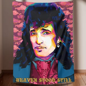 Könnte beinhalten: Eine farbenfrohe Pop-Art-Tapisserie mit einem Porträt einer Person mit langen dunklen Haaren und einem pink-, blau- und gelb gemusterten Hemd. Der Text "Heaven Stood Still" ist am unteren Rand der Tapisserie gedruckt.
