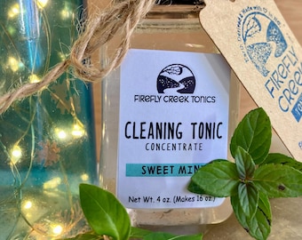 May include: A clear glass bottle with a black lid and a brown paper tag tied around it. The bottle contains a clear liquid and has a label that reads "FIREFLY CREEK TONICS CLEANING TONIC CONCENTRATE SWEET MINT Net Wt. 4 oz. (Makes 16 oz.)". There is a sprig of green mint leaves next to the bottle.