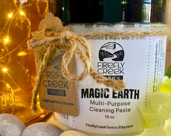 May include: A white jar with a black lid and a brown paper label. The label says "Firefly Creek Tonics" and "Magic Earth Multi-Purpose Cleaning Paste 16 oz. FireflyCreekTonics.Etsy.com". The jar is sitting on a white rock.