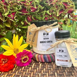 May include: Two glass jars with white labels and brown twine. The larger jar contains a white cleaning paste with the text "Firefly Creek Tonics Magic Earth Multi-Purpose Cleaning Paste 16 oz". The smaller jar contains a clear liquid with the text "Firefly Creek Tonics Cleaning Tonic Concentrate Woodlands Net Wt. 4 oz. (Makes 16 oz)".