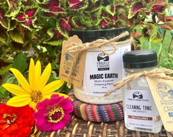 May include: Two glass jars with white labels and brown twine. The larger jar contains a white cleaning paste with the text "Firefly Creek Tonics Magic Earth Multi-Purpose Cleaning Paste 16 oz". The smaller jar contains a clear liquid with the text "Firefly Creek Tonics Cleaning Tonic Concentrate Woodlands Net Wt. 4 oz. (Makes 16 oz)".