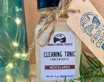 May include: A clear glass bottle with a black lid and a brown paper tag tied around the neck. The bottle contains a light yellow liquid and is labeled "FIREFLY CREEK TONICS CLEANING TONIC CONCENTRATE WOODLANDS Net Wt. 4 oz. (Makes 16 oz.)".