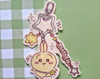 May include: A yellow bunny keyring with a star-shaped charm and the word "USAGI" on a charm. The keyring has a silver clasp and is decorated with small stars. The background is a green and white checkered pattern.