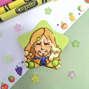 May include: A star-shaped pin featuring a cartoon illustration of a person with long blonde hair, a goatee, and a blue and tan outfit. The background of the pin is a light green colour with a subtle dot pattern, and the character is surrounded by small green spheres.