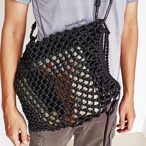 May include: A black paracord net bag featuring a thick braided shoulder strap attached with a black carabiner. The bag has a square-knot mesh pattern and displays the text kenatastudio on the strap and near the top edge of the bag structure.