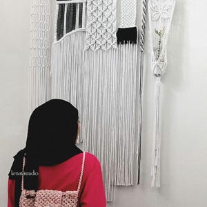 May include: A macrame wall hanging with geometric and fringed designs in white and black. A small, pink crocheted backpack is in the foreground. The wall hanging is suspended from a wooden branch.