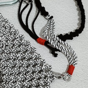 May include: A macrame plant hanger with a black and white pattern, a black rope, and a silver carabiner. The hanger has red accents and the text "kenatastudio".