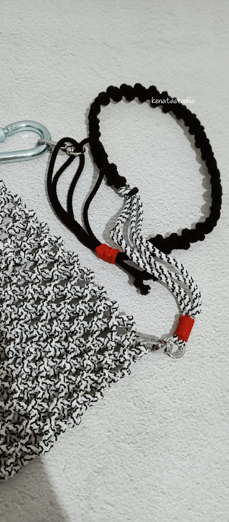 May include: A macrame plant hanger with a black and white pattern, a black rope, and a silver carabiner. The hanger has red accents and the text "kenatastudio".
