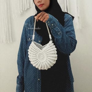 May include: A white, crocheted handbag with a seashell design. The bag has a circular shape with a beaded strap. The text "kenatastudio" is visible on the image. The bag is held by a person wearing a denim shirt.