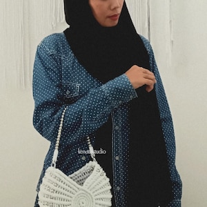 May include: A white, crocheted seashell-shaped handbag with a pearl strap. The bag is held against a blue denim shirt with white polka dots. The text "kenat studio" is visible on the shirt.