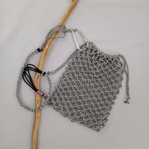 May include: A small, hand-knitted bag in black and white, with a drawstring closure and a silver carabiner clip. The bag has a matching black and white strap with a black cord and pink accents. The bag is designed to be carried.