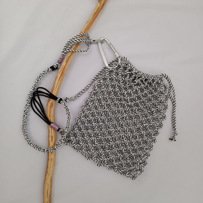 May include: A small, hand-knitted bag in black and white, with a drawstring closure and a silver carabiner clip. The bag has a matching black and white strap with a black cord and pink accents. The bag is designed to be carried.