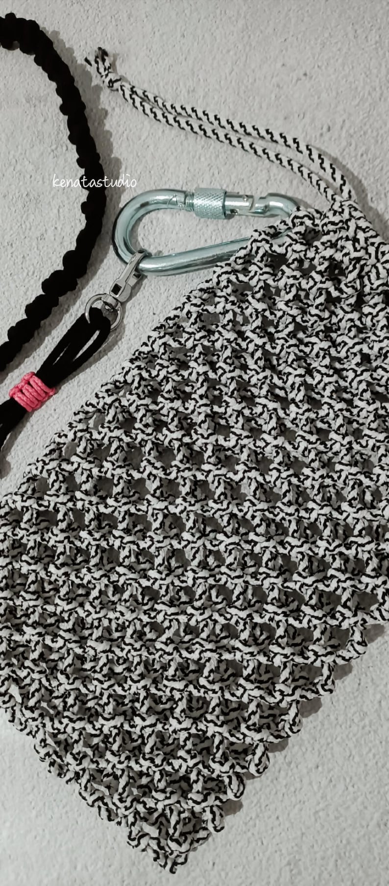 May include: A macrame phone case in black and white, attached to a silver carabiner clip. A black cord with a pink accent and a black cord are also visible. The brand name "kenatastudio" is in the upper left corner.
