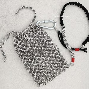 May include: A black and white macrame bag with a drawstring closure, connected to a black and white rope leash with a silver carabiner. The leash has red accents. The bag and leash are on a white surface.