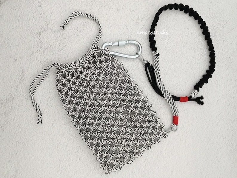 May include: A black and white macrame bag with a drawstring closure, connected to a black and white rope leash with a silver carabiner. The leash has red accents. The bag and leash are on a white surface.