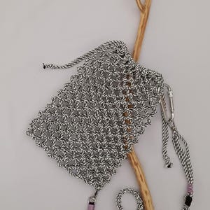 May include: A small, woven bag in black and white with a drawstring closure. The bag has a long strap with a silver clasp and a black and pink accent. A wooden branch is in the background.